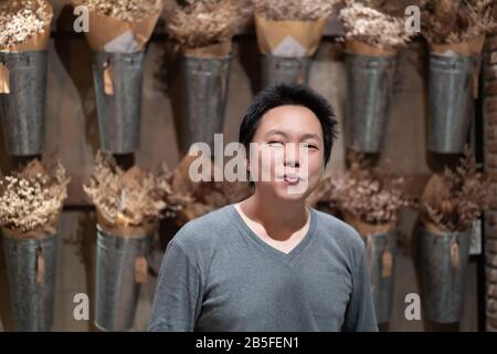 L'uomo asiatico felice dell'adulto sorride alla macchina fotografica davanti al fiore asciutto verticale in vaso di alluminio sulla parete. Foto Stock