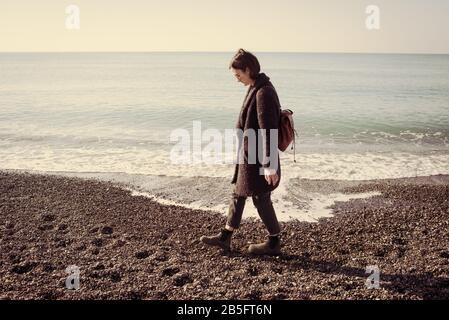 Donna modello hippster indossando occhiali da sole e cappotto invernale a piedi su una spiaggia di ghiaia con vuoto mare calmo dietro. Effetto vintage opaco. Foto Stock