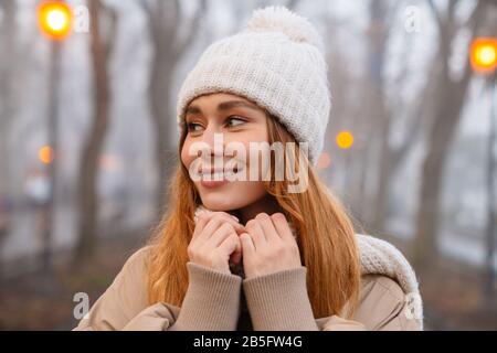 Attraente giovane ragazza sorridente che indossa abiti invernali in piedi al parco in serata Foto Stock