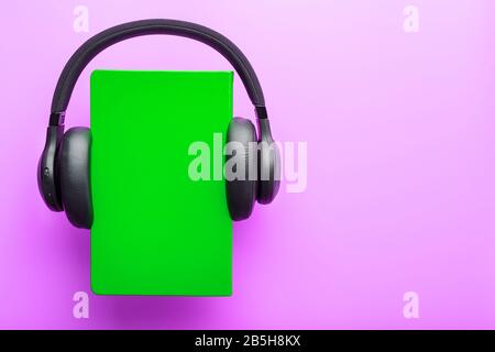 Le cuffie sono indossate su un libro in una copertina rigida verde su uno sfondo lilla, vista dall'alto. Il concetto Di Audiolibro e formazione A Distanza, e-learning. Copia Foto Stock
