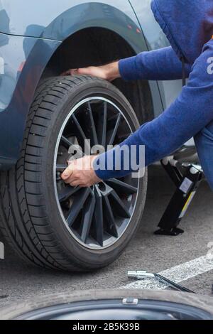 Un uomo in un giubbotto di sicurezza arancione cambia una gomma piatta su una strada. Primo piano le mani mans al volante di una vettura rotta. Sostituzione di una ruota con l'attrezzo Foto Stock