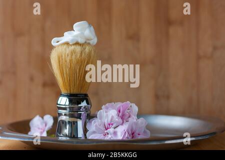 Spazzola da barba con fofarm sulla parte superiore e fiori di geranio di colore rosa tenero sul lato di vaso in acciaio invecchiato su sfondo di legno. Cura della pelle per gli uomini. Plac Foto Stock