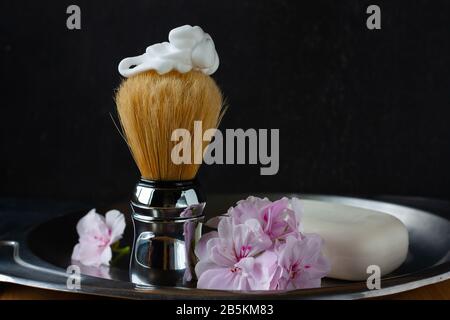 Spazzola da barba con fofarm sulla parte superiore e fiori di geranio di colore rosa tenero e sapone bianco sul lato di vaso in acciaio invecchiato su sfondo nero. Cura della pelle Foto Stock