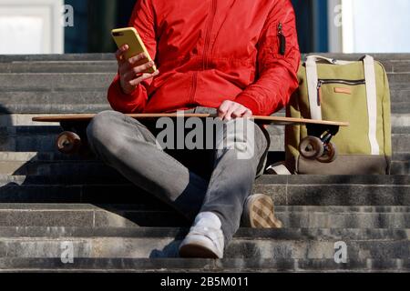 Elegante longboarder uomo in abiti casual con il suo smartphone, appoggiato sui gradini, seduto con longboard/skateboard all'aperto, vista frontale, corto Foto Stock