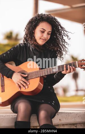 Ragazza brunetta con capelli neri ricci che indossa nero suona la chitarra classica in un parco cittadino Foto Stock