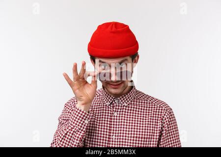 Primo piano ritratto di sospetto giovane ragazzo divertente in berretto rosso e camicia controllata, prendere occhiali da sole e guardare da sotto la fronte come pettegolezzi Foto Stock