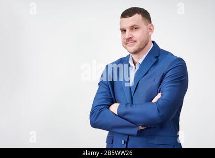 Giovane uomo d'affari di successo con le braccia incrociate che indossano vestito blu con un sorriso genuino grazioso che posano contro lo sfondo grigio con lo spazio della copia Foto Stock