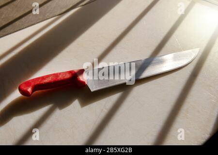 Coltello da macellaio con manico rosso, visto in primo piano, seduto sulla superficie bianca del tavolo da taglio in ombra dalle tende della finestra Foto Stock