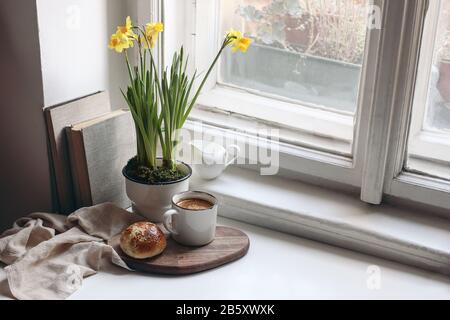 Accogliente primavera di Pasqua ancora vita scena. Tazza di caffè, dolce bun, libri, tagliere in legno, caraffa latte e vaso di fiori su vecchio davanzale. Floreale Foto Stock