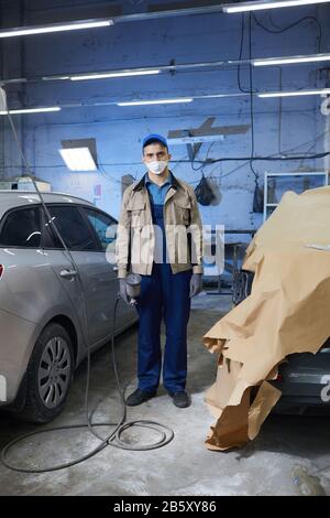 Scatto verticale completo di un pittore di auto irriconoscibile indossando abiti protettivi e maschera sul viso in piedi in officina guardando la fotocamera Foto Stock