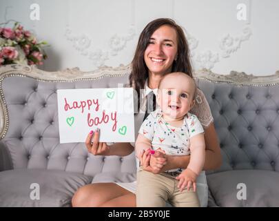 Giovane donna e figlio che tiene la carta nelle braccia. Piccolo bambino senza riposo seduto su un divano in un elegante interno. Concetto di cura del Padre Foto Stock