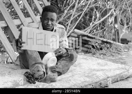 Giovane uomo africano senza tetto con segno di cartone che chiede aiuto in bianco e nero Foto Stock