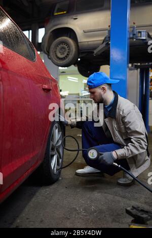 Colpo di vista laterale verticale di un riparatore professionista seduto davanti alla ruota dell'automobile usando il manometro degli pneumatici Foto Stock