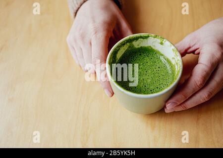 una tazza di latte da tè verde matcha in una tazza di ceramica verde e le mani delle donne su un tavolo di sfondo di legno. Spazio di copia. Foto Stock