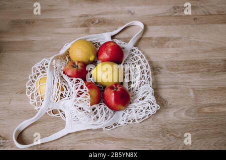 Borsa a mano in rete bianca con mele rosse e limoni gialli su sfondo in legno. Vista superiore, spazio di copia. Concetto ecologico. Concetto di zero sprechi. Foto Stock