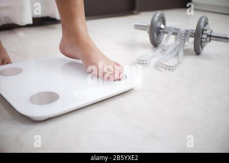 Immagine ritagliata dei piedi della donna in piedi su scale di pesatura, un metro a nastro e dumbbell in primo piano Foto Stock