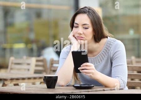 Annoiata ragazza guardando deluso per il suo smartphone su una terrazza caffè Foto Stock