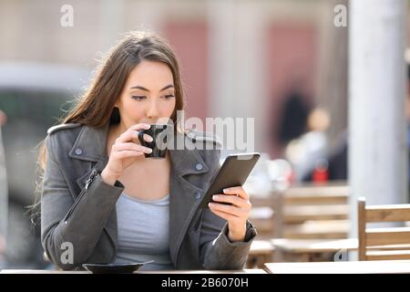 Ritratto di una giovane donna che beve una tazza di caffè controllando il suo smartphone su una terrazza caffetteria Foto Stock