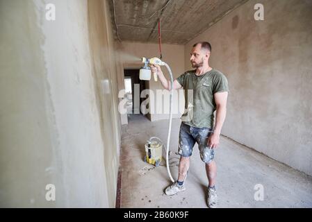 Ritratto a tutta lunghezza del costruttore maschio che lavora con aerografo, riparazione della casa. Grande volume di lavoro è visibile sullo sfondo. L'uomo con la barba è vestito con vestiti spalmati di vernice. Concetto di riparazione Foto Stock