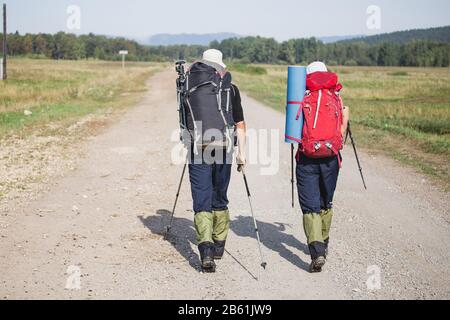 Vista sul retro di un paio di escursionisti con zaini che camminano lungo una strada rurale empry Foto Stock