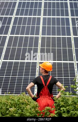 Vista posteriore del lavoratore in uniforme arancione e casco in piedi sullo sfondo di grandi pannelli solari piantagione. Costruzione domestica di stazione solare. Scatto verticale. Generazione di energia ecologica verde Foto Stock