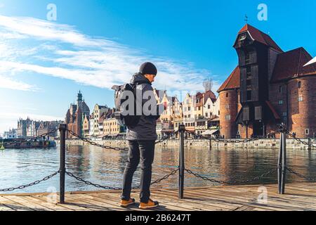 Tema del turismo in Polonia. Viaggiare a Danzica. Un viaggiatore maschio usa un telefono sull'argine del fiume Mottawa in mezzo all'attrazione principale, un Foto Stock