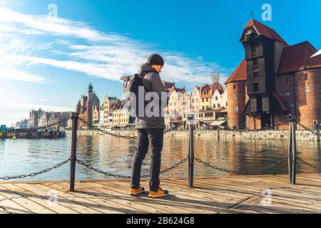 Tema del turismo in Polonia. Viaggiare a Danzica. Un viaggiatore maschio usa un telefono sull'argine del fiume Mottawa in mezzo all'attrazione principale, un Foto Stock