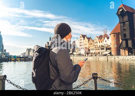 Tema del turismo in Polonia. Viaggiare a Danzica. Un viaggiatore maschio usa un telefono sull'argine del fiume Mottawa in mezzo all'attrazione principale, un Foto Stock