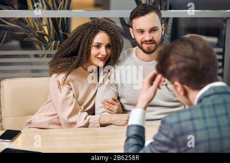 elegante uomo d'affari di stile che ha una riunione con una coppia seduta insieme ad un tavolo nell'ufficio, la famiglia che va prendere il prestito, affittando un appartamento, libro Foto Stock