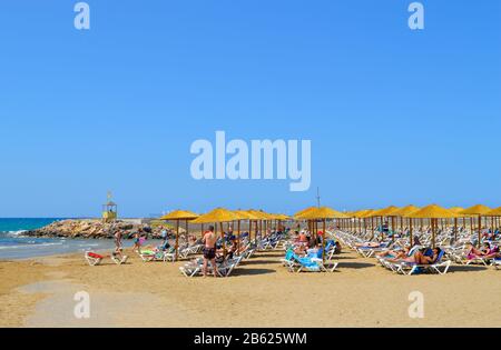 Turisti prendere il sole sotto ombrelloni sulla spiaggia di Gouves a Creta, la più grande e più popolata dell'isola greca Foto Stock