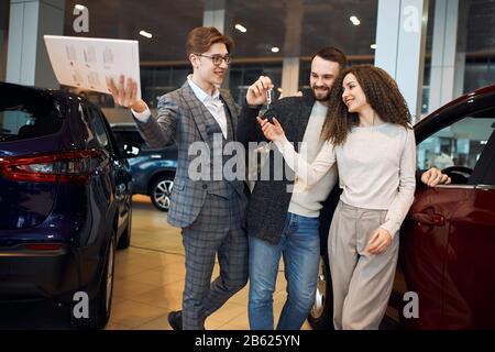 il commerciante professionista riuscito di cheeful sta manando le chiavi auto ad una coppia giovane, sono eccitati, le automobili che si levano in piedi in background, contrattare, sogno viene Foto Stock