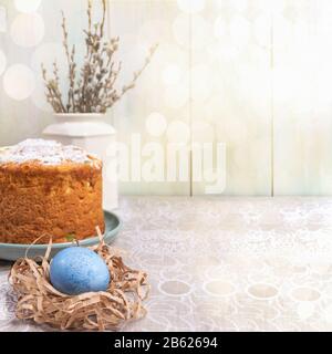 Pasqua sfondo festoso con torta di Pasqua, uovo blu in nido di carta e salice rami in vaso bianco su tovaglia di lino contro legno dipinto di luce Foto Stock