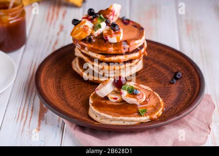 Deliziose e lussureggianti frittelle con frutta fresca e frutti di bosco, versate con caramello salato. Menta, tè, succo d'arancia, latte sul tavolo. Colazione gustosa Foto Stock