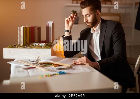 giovane e bello, pensoso e pensoso, bruna bearded in elegante abito nero con una matita in mano e pensando allo sviluppo del vicinato Foto Stock