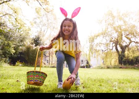 Ritratto Di Ragazza Che Indossa Orecchie Di Coniglio Trovare Uovo Di Cioccolato Sulla Caccia All'Uovo Di Pasqua In Giardino Foto Stock