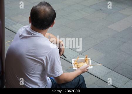 Uomo asiatico seduto per strada, tenendo un granchio fritto profondo su uno spiedino, pronto per mangiare. Shanghai, Cina. Foto Stock