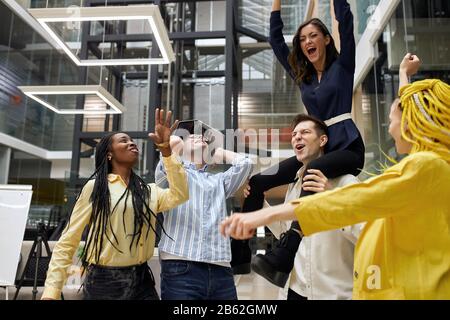 team multirazziale entusiasta di divertirsi a cavalcare in ufficio, eccitato dipendenti diversi ridere godendo di attività divertente sul posto di lavoro. follia, follia Foto Stock