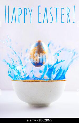 Un uovo di Pasqua in perla dipinta in blu e oro vola in una ciotola di vernice blu. Lettere Felice Pasqua. Biglietto d'auguri per la vacanza. Foto Stock