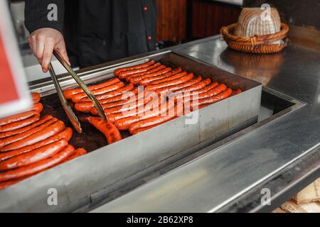 Primo piano di mano d'uomo, cucinando salsicce speziate sulla griglia calda piatta in strada Foto Stock