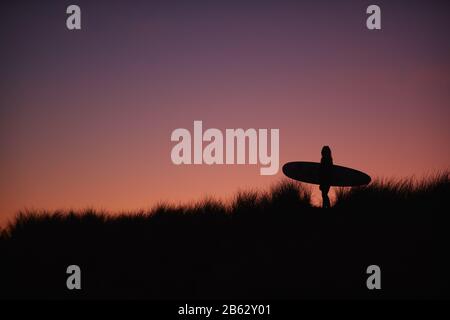 Silhouette Di Surfer Femminile Che Trasporta Il Surfboard Attraverso Le Dune Contro Il Sole Del Setting Foto Stock