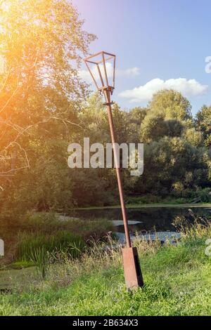 Paesaggio estivo. Nel vecchio parco sullo sfondo di fitti alberi verdi e la superficie scura dello stagno sorge arrugginita, pendente, rotto lanterna elettrica in soli Foto Stock