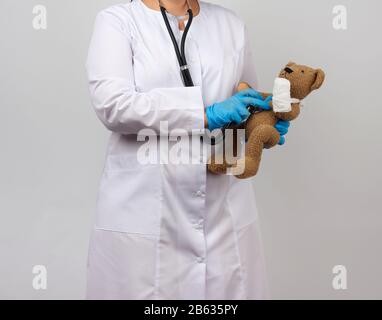 il medico femminile tiene l'orsacchiotto marrone con la zampa bendata in bendaggio bianco e ascolta il giocattolo con stetoscopio, concetto di pediatria e trattamento degli animali Foto Stock