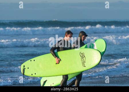 Due surfisti che portano le tavole da surf noleggiate a piedi fuori dal mare a Fistral a Newquay in Cornovaglia. Foto Stock