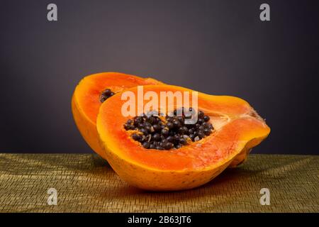 Brillante e vivace frutto di papaia arancione affettato a metà su una superficie color oro con semi neri che si accumulano contro uno sfondo grigio scuro Foto Stock