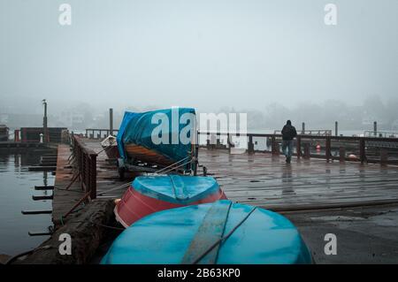 Un uomo cammina verso la fine di un molo di lavoro a Gloucester Massachusetts in una mattina fredda e piovosa. Quattro barche di legno colorate ancorate a secco Foto Stock