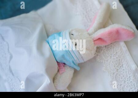 Coniglio in una maschera medica protettiva si trova in una culla su un letto bianco. Il giocattolo si ammalò. Medico. Protezione da virus, influenza, coronavirus f Foto Stock