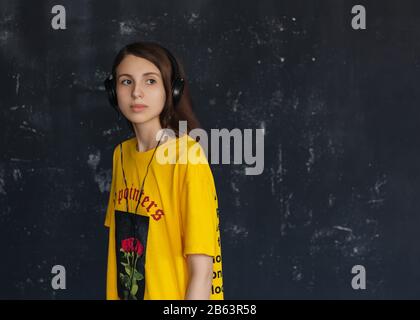 La ragazza meditativa in una lunga t-shirt gialla sta ascoltando musica sulle cuffie Foto Stock