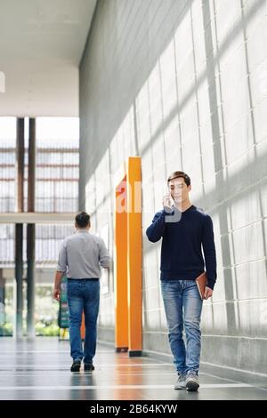 Bel giovane specialista IT che cammina nel corridoio dell'ufficio e parla al telefono Foto Stock