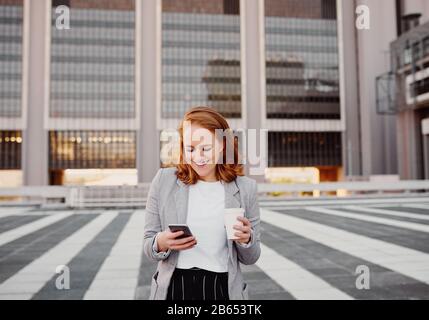 Una donna di successo che utilizza uno smartphone in piedi all'aperto con una tazza da caffè da asporto Foto Stock