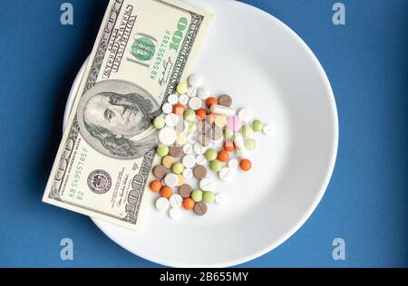 I farmaci costano denaro. Drugs and dolls.White Plate con pillole e capsule multicolore e una fattura da 100 dollari su un classico sfondo blu. Pillole Foto Stock